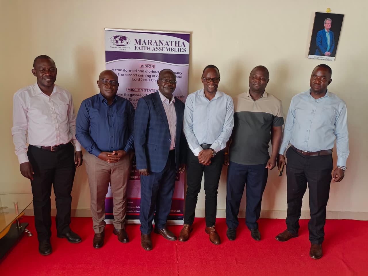 Six men standing in front of a Maranatha Faith Assemblies banner at a meeting
